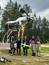 Fem fiskare och en kapten står framför en stor blank älgstaty i skogen med svenska flaggor.