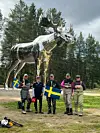 Fem fiskare och en kapten står framför en stor blank älgstaty i skogen med svenska flaggor.
