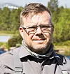 Tony Forsberg med glasögon och grå skjorta framför träd och vatten.
