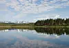 Lugn sjö med skog, röda stugor och snötäckta fjäll i bakgrunden.